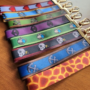 May include: A collection of colorful keychains with various patterns. Designs include hearts, fish, crossed swords, skulls, anchors, and a giraffe-like pattern. Each keychain has a gold-colored clasp.