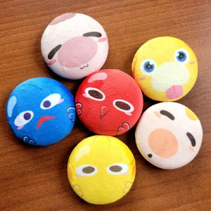 May include: A collection of six plush, round, emoji-style toys with various facial expressions. The toys are in shades of blue, red, yellow, and white, with black and pink accents. The toys are arranged on a wooden surface.