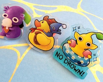 May include: Three whimsical, clear acrylic pins with cartoon characters. One purple creature holds an ice cream cone. Another features a yellow duck with a blue creature. The third has a yellow character in a floatie, with the text "NO DROWN!"