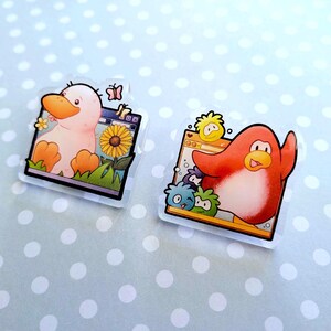 May include: Two clear acrylic pins featuring cartoon characters. One pin shows a duck with an orange beak and feet, a sunflower, and a butterfly. The other pin features a red penguin with a yellow beak and a few small, colorful creatures.