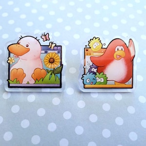 May include: Two clear acrylic pins featuring cartoon characters. One pin shows a pink duck with an orange beak and feet, a sunflower, and butterflies. The other pin depicts a red penguin with a raised wing and other cartoon characters.