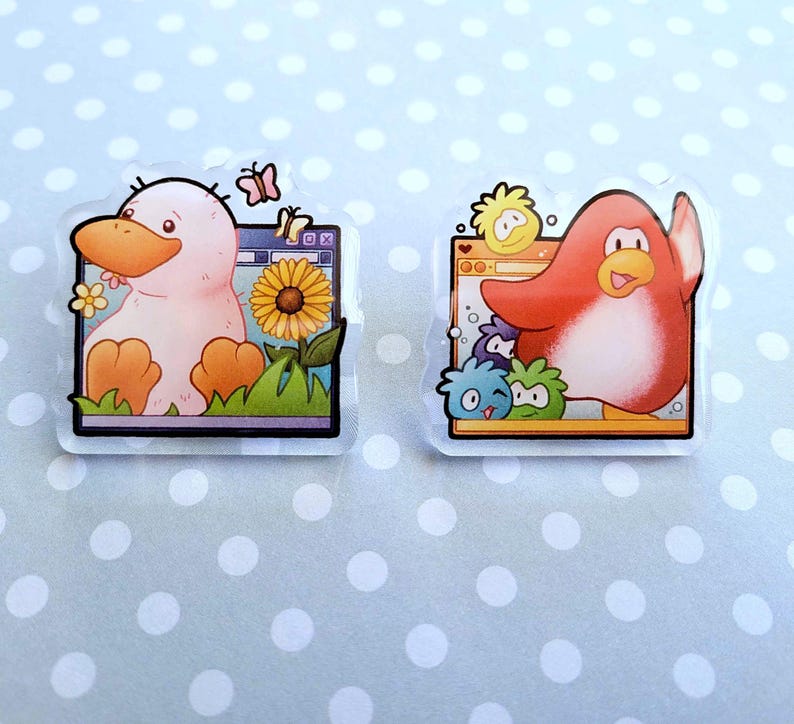 May include: Two clear acrylic pins featuring cartoon characters. One pin shows a pink duck with an orange beak and feet, a sunflower, and butterflies. The other pin depicts a red penguin with a raised wing and other cartoon characters.