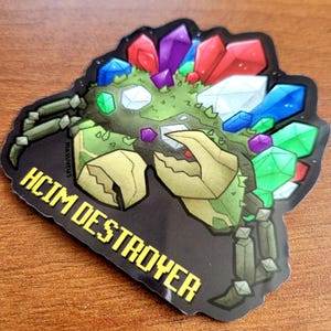 May include: A sticker featuring a crab-like creature with a green body, tan claws, and legs. The crab is adorned with colorful, gem-like crystals in red, purple, blue, and green. The text "HCIM DESTROYER" is in yellow block letters.