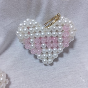 Peut inclure: Un porte-clés en forme de cœur composé de perles blanches et roses. Le porte-clés a un anneau en métal doré.