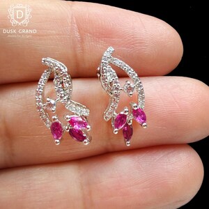 May include: A pair of silver-toned stud earrings featuring a floral design. Each earring has a curved, openwork shape, encrusted with small, clear crystals. The design includes three vibrant, oval-cut, ruby-red gemstones. The earrings are displayed on a hand.