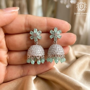 May include: A pair of silver-toned jhumka earrings featuring a flower-shaped stud with light green oval stones and a central clear stone. The bell-shaped drop is covered in small clear crystals and finished with dangling light green beads.