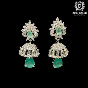 May include: A pair of silver-toned dangle earrings with a floral design at the top and a bell-shaped bottom. The earrings feature clear and emerald-green gemstones. The Dusk Grand logo is in the upper right corner.