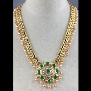 May include: A gold-toned necklace with a central pendant and matching chain. The necklace features emerald green and clear gemstones, with a pearl-like trim. The pendant has a large green stone at its center.