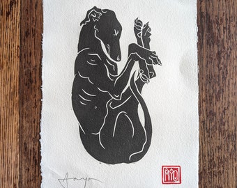 May include: A black and white linocut print of a dog curled up. The dog is in a relaxed pose, with its legs crossed. The print is on textured paper, with the artist's signature and a red stamp in the corner.