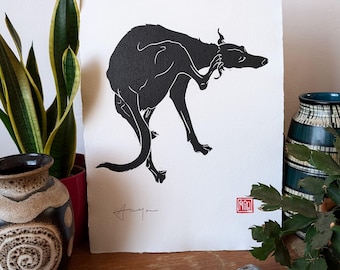 May include: Black and white linocut print of a dog scratching its ear. The print is on white paper and has a red stamp in the bottom right corner.