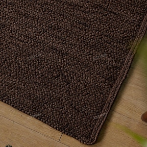May include: A rectangular area rug featuring a dark brown, textured, woven surface with a thick, stitched border. The material displays a dense, knotted pattern throughout, providing a uniform appearance across the entire visible surface area.