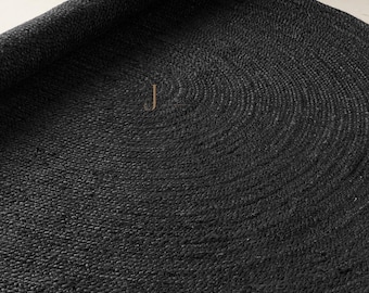 May include: A round, black, braided area rug with a textured surface. The rug is partially rolled up, revealing a circular shape and a consistent, woven pattern throughout the entire piece. The material appears to be a natural, thick fiber.