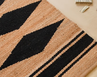 Puede incluir: Una alfombra rectangular de pasillo hecha de fibra de yute natural en tono bronceado, con un patrón repetitivo de grandes diamantes negros y dos franjas horizontales negras en el extremo. La alfombra tiene una textura tejida y plana.