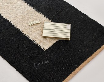 Puede incluir: Una alfombra rectangular con una ancha franja central de color crema flanqueada por dos secciones negras sólidas. La alfombra tiene un borde de yute natural de color tostado en los extremos y una superficie tejida texturizada. Una pequeña muestra de tela a rayas doblada descansa sobre ella.