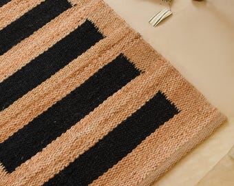 Puede incluir: Una alfombra rectangular tipo pasillero con una textura tejida de yute natural en tono bronceado y rayas horizontales negras. La alfombra presenta una construcción de fibras orgánicas gruesas sobre una superficie clara con el logo Jute Hub.