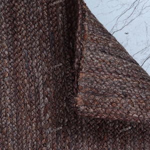 May include: A dark brown rectangular rug featuring a thick, braided jute texture. The rug is folded at one corner, revealing the dense, woven construction of the natural fibers. The surface displays a consistent, deep brown color throughout the weave.