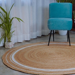 Könnte beinhalten: Ein runder Jute-Teppich in Naturbeige mit weißem Rand. Ein türkisfarbener Stuhl mit schwarzen Beinen steht auf dem Teppich. Der Teppich hat einen Durchmesser von etwa 152 cm. Pflanzen und Vorhänge sind im Hintergrund zu sehen.
