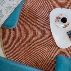 Könnte beinhalten: Draufsicht auf ein Wohnzimmer mit einem runden, rot-beige gestreiften Teppich. Ein weißer, dreieckiger Couchtisch trägt eine Kaffeetasse und ein Buch mit dem Titel "Think and Grow Rich". Türkisfarbene Möbel und eine gemusterte Decke ergänzen die Szene.