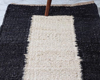 May include: A rectangular rug with a central cream-colored stripe bordered by black. The rug has a textured appearance and a natural fiber look. The rug is placed on a light-colored floor.