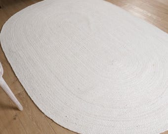 Puede incluir: Una alfombra ovalada de yute blanquecina. La alfombra está hecha de fibras tejidas y tiene una superficie texturizada. La alfombra está colocada sobre un suelo de madera de color claro. La forma de la alfombra es un óvalo grande.