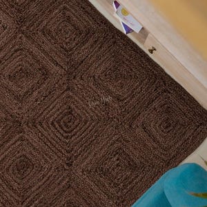 Könnte beinhalten: Ein dunkelbrauner Teppich mit einem sich wiederholenden quadratischen Muster und einer strukturierten, gewebten Oberfläche. Der Teppich liegt auf dem Boden und ist teilweise von einem runden, hellen Beistelltisch mit Tasse und einem blauen Möbelstück bedeckt.