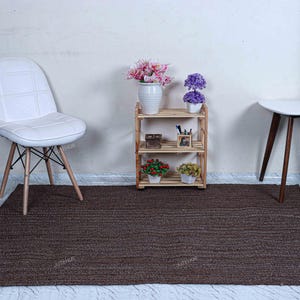May include: A rectangular dark brown area rug with a textured, woven surface. The rug is positioned on a light-colored floor in front of a white chair, a three-tiered wooden shelf containing small potted plants, and a small white side table.