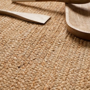 May include: Close-up of a woven jute rug with a natural, light brown color. Wooden kitchen utensils, including a spatula and a serving spoon, are partially visible. The rug's texture and the wooden items suggest a rustic, eco-friendly theme.