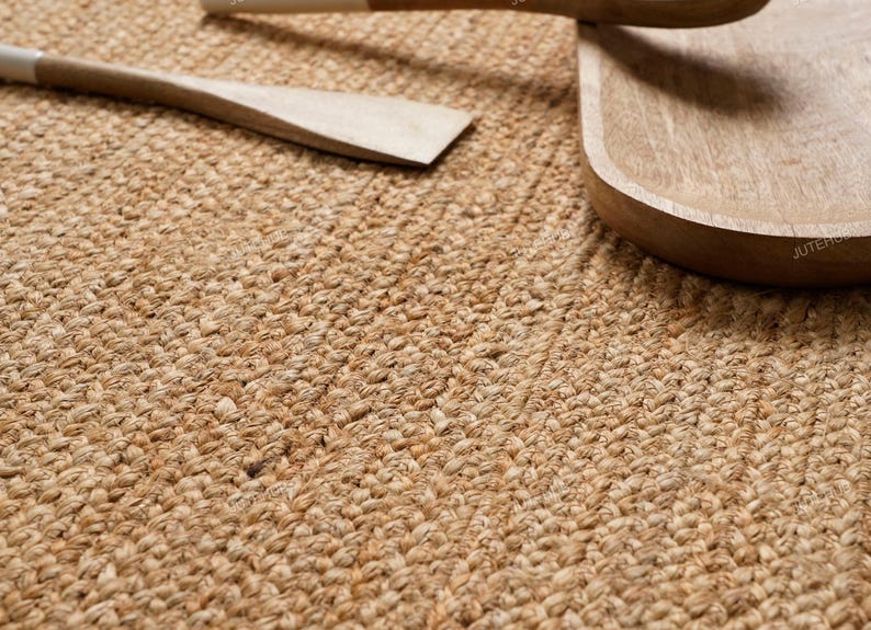 May include: Close-up of a woven jute rug with a natural, light brown color. Wooden kitchen utensils, including a spatula and a serving spoon, are partially visible. The rug's texture and the wooden items suggest a rustic, eco-friendly theme.