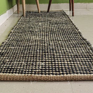 May include: A rectangular runner rug featuring a woven pattern of alternating black and off-white circular fibres. The rug has a thick, natural tan-coloured braided border along the short edge, creating a textured surface with a repeating grid design.