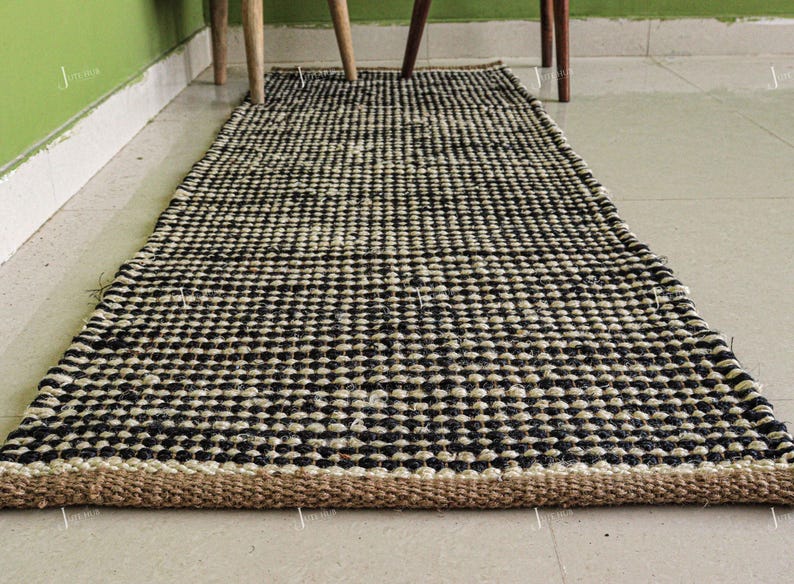 May include: A rectangular runner rug featuring a woven pattern of alternating black and off-white circular fibres. The rug has a thick, natural tan-coloured braided border along the short edge, creating a textured surface with a repeating grid design.