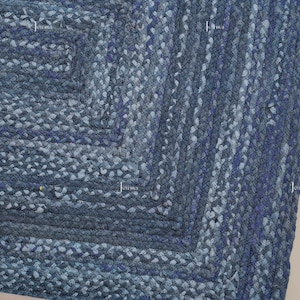 May include: A close-up of a blue braided rug with a square design. The rug is made of thick, textured material in shades of blue and grey. The braided pattern creates a geometric design, suitable for home decor.