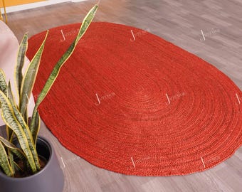 Könnte beinhalten: Ovaler, handgewebter Jute-Teppich in einem leuchtenden Rot-Orange-Ton. Das Design des Teppichs weist konzentrische Kreise auf, die eine strukturierte Oberfläche erzeugen. Eine Topf-Bogenhanf-Pflanze mit grünen und gelben Blättern befindet sich im Vordergrund.