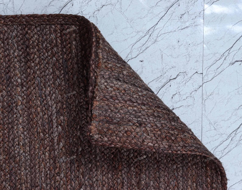 May include: A dark brown rectangular rug featuring a thick, braided jute texture. The rug is folded at one corner, revealing the dense, woven construction of the natural fibers. The surface displays a consistent, deep brown color throughout the weave.