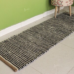 May include: A rectangular runner rug featuring a woven texture with a repeating pattern of black and off-white horizontal stripes. The rug has a natural tan jute border along the short ends and a thick, braided construction throughout the surface.