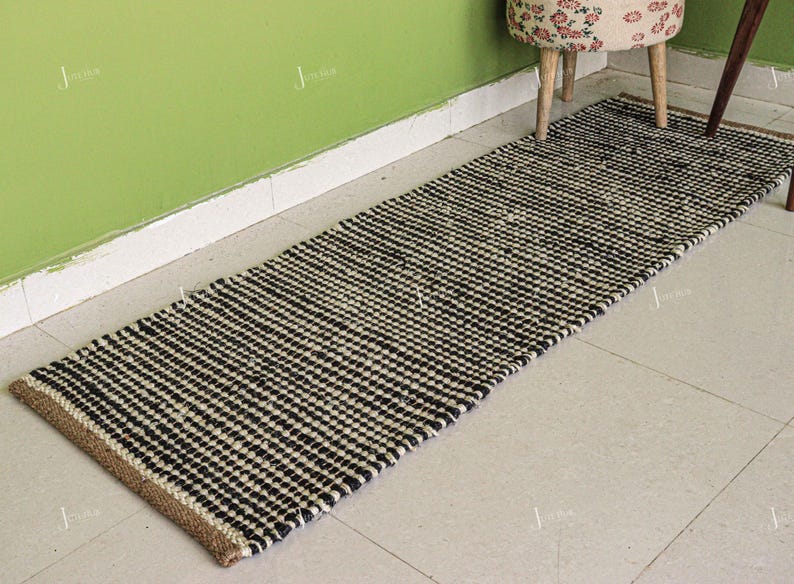 May include: A rectangular runner rug featuring a woven texture with a repeating pattern of black and off-white horizontal stripes. The rug has a natural tan jute border along the short ends and a thick, braided construction throughout the surface.