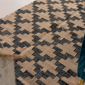 May include: A rectangular jute rug with a geometric pattern in black and beige. The rug's design features a repeating zigzag motif. A teal armchair is visible in the corner. The rug is likely used for home decor.