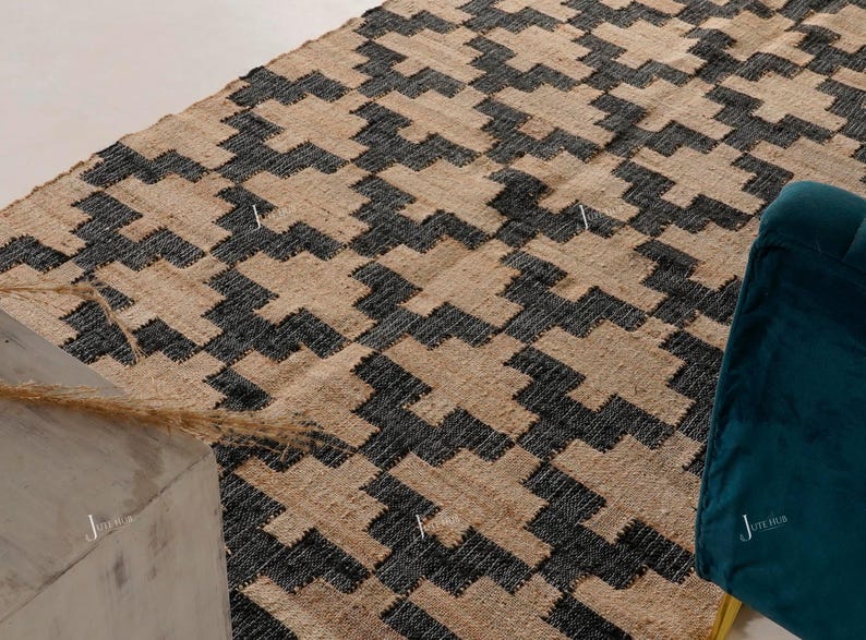 May include: A rectangular jute rug with a geometric pattern in black and beige. The rug's design features a repeating zigzag motif. A teal armchair is visible in the corner. The rug is likely used for home decor.