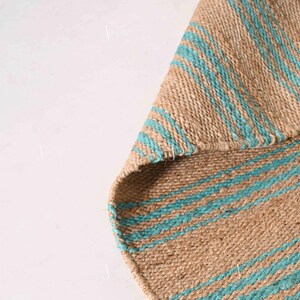 Op de afbeelding: Een geweven jute vloerkleed met afwisselende strepen in natuurlijk bruin en turquoise blauw. De textuur van het vloerkleed is zichtbaar, met een opgevouwen hoek die de onderkant laat zien. Geschikt voor gebruik binnenshuis.