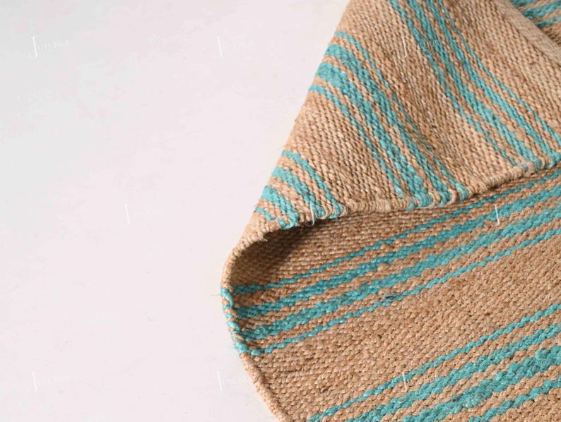Op de afbeelding: Een geweven jute vloerkleed met afwisselende strepen in natuurlijk bruin en turquoise blauw. De textuur van het vloerkleed is zichtbaar, met een opgevouwen hoek die de onderkant laat zien. Geschikt voor gebruik binnenshuis.