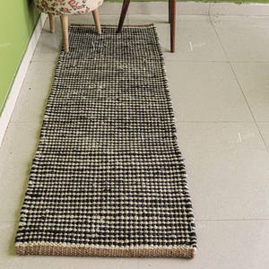 May include: A rectangular runner rug featuring a woven texture with a repeating pattern of alternating black and natural beige horizontal stripes. The rug has a thick, knotted construction and a flat, low-profile design laid out on a tiled floor.