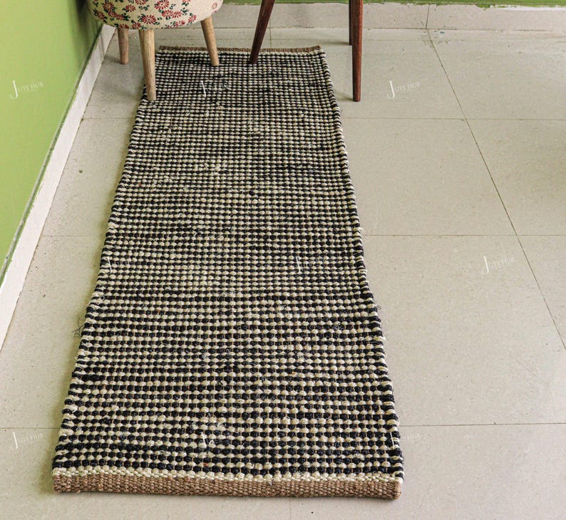 May include: A rectangular runner rug featuring a woven texture with a repeating pattern of alternating black and natural beige horizontal stripes. The rug has a thick, knotted construction and a flat, low-profile design laid out on a tiled floor.