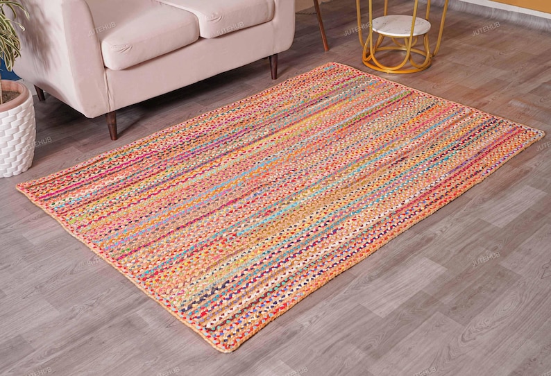May include: A rectangular, multi-colored braided rug with a striped pattern. The rug features a mix of orange, pink, yellow, and blue hues. It is placed on a wooden floor, and a beige sofa is partially visible in the background.