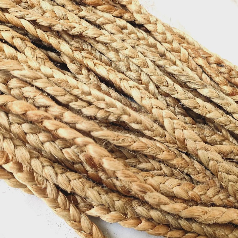 May include: Close-up of several strands of braided jute rope. The natural fibers are a light brown color, with variations in tone. The braids are tightly woven, creating a textured surface. The rope appears to be new and unused.
