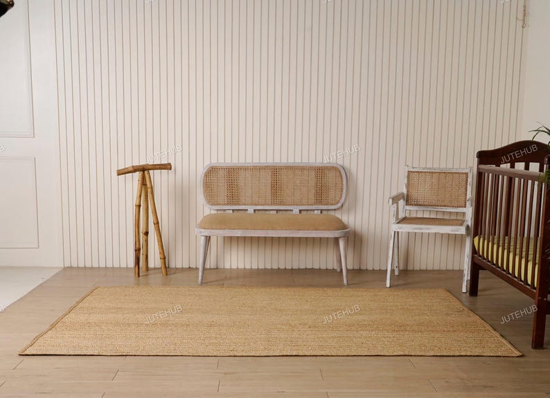 May include: A natural fiber jute rug is laid on a light wood floor. A wooden bench with a woven back and seat, a matching chair, and a crib are in the background. A bamboo tripod stand is on the left.
