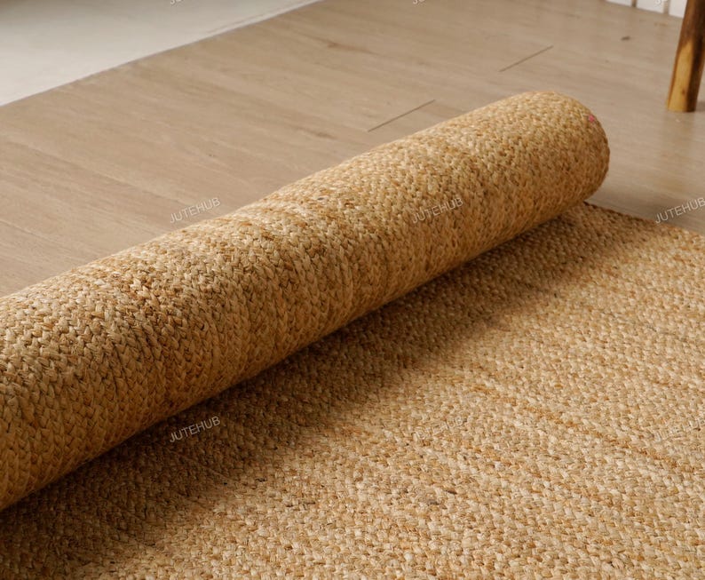 May include: A rolled-up, natural-toned jute rug rests on a light-colored wooden floor. The rug's texture is visible, showcasing its woven construction. The unrolled portion of the rug lies flat, revealing the full weave pattern. The rug is a neutral color, suitable for various interior styles.