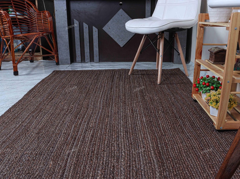 Op de afbeelding: Een rechthoekig vloerkleed met een donkerbruin, gevlochten en getextureerd oppervlak. Het kleed ligt op een lichte vloer en strekt zich uit van de voorgrond naar het midden, onder een witte stoel en bij een houten rek.