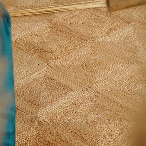 May include: Close-up of a natural jute rug with a geometric diamond pattern. The rug is a warm beige color, with a textured surface. The rug is made of woven jute fibers, and is suitable for indoor use.