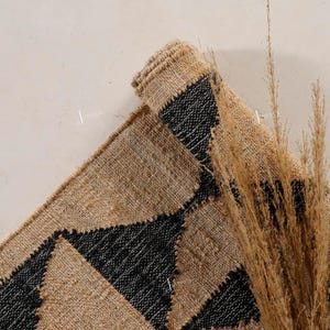 May include: A rolled jute rug featuring a geometric design of black triangles on a natural tan backdrop. The image includes dried ornamental grass, providing a textural and colourful contrast to the rug.