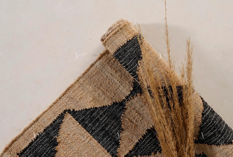 May include: A rolled jute rug featuring a geometric design of black triangles on a natural tan backdrop. The image includes dried ornamental grass, providing a textural and colourful contrast to the rug.