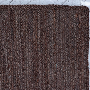 May include: A rectangular area rug featuring a dark brown, braided jute construction. The rug displays a consistent, tight weave pattern with a thick, reinforced border edge that frames the textured surface, showcasing natural fiber variations.